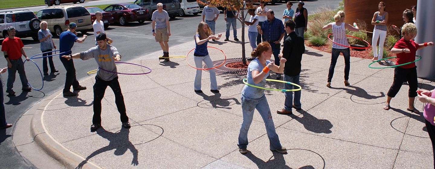 Landscape Structures employees competing in hula hoop challenge outside office building on a sunny day.