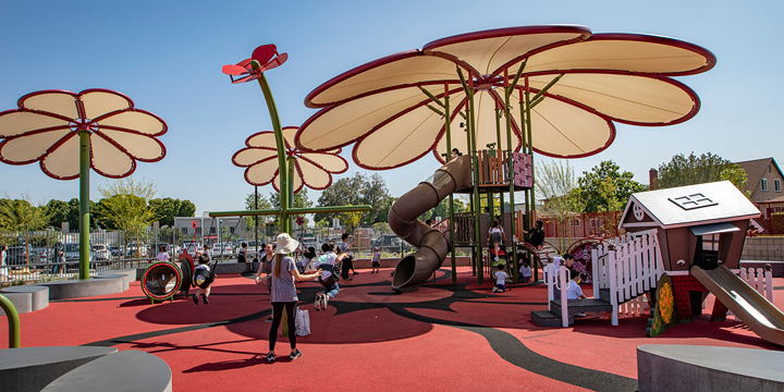 A playground with red, brown and pink colors has three white flower-shaped shades overhead and one pink flower-motif post topper. 