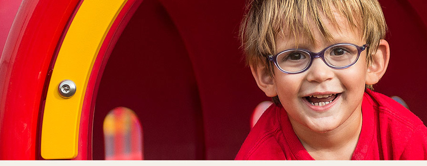 Young boy with glasses smiles wall crawling out of red playground crawl tunnel.