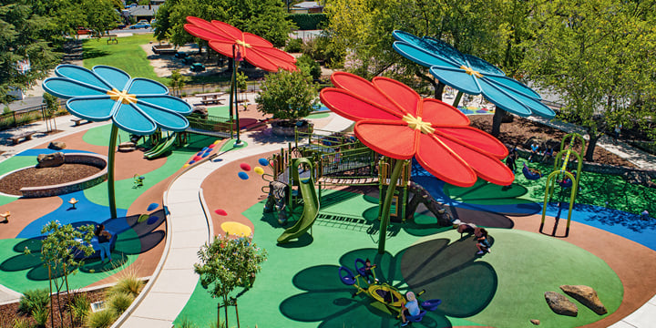 Flower-shaped shade structures in red and turquoise cover a playground with green surfacing. 
