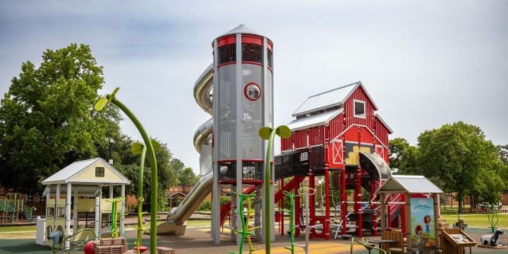 A farm-themed playground with a yellow playhouse on the left, a silver silo tower in the middle and a red barn climber to the right. 