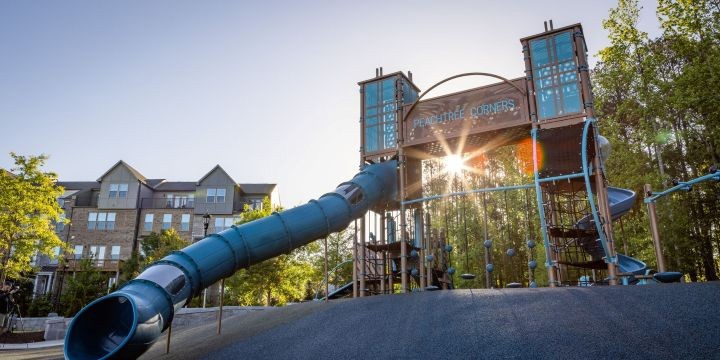 Looking up at two playground towers connected by a tunnel with "Peachtree Corners" written on it. A blue twisty slide descends to the left. 