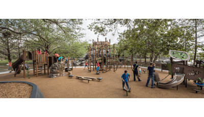 Families enjoy this nature-inspired playground. A father helps his toddler down a PlayShaper slide. A group of older kids play on the Geoplex net structure.