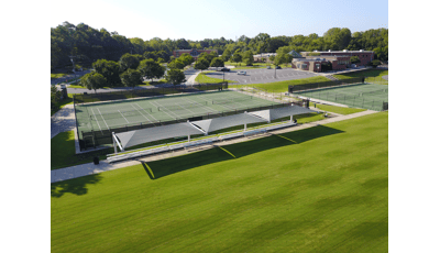 Elevated view of a lush green field with a sidewalk containing rows of bleachers covered by three large rectangular shade systems and a fenced in location behind with three tennis courts. 