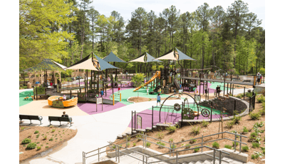 Filled with bridges and ramps, the playground also has tan and green multi-panel shade structures. This is a large playground with orange slides and surrounded by tall green trees.