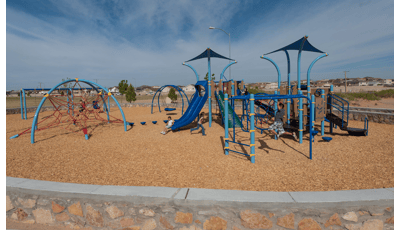 Children play on a park playground with different shades of blue posts, overhead shades, and climbers all surrounded by a neighborhood community. 